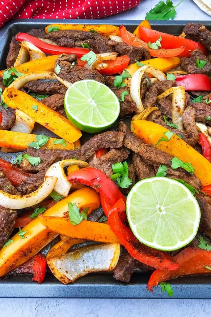 Easy steak fajitas fill a sheet pan with seasoned strips of beef. Roasted red and orange bell peppers and onions are topped with fresh cilantro and bright green lime halves for a colorful one pan meal.