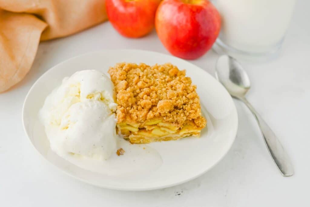 Apple crumble bar with tender cinnamon apple layers and a golden crumb topping served with melting vanilla ice cream. Two red apples and a glass of milk complete the cozy fall dessert setting.