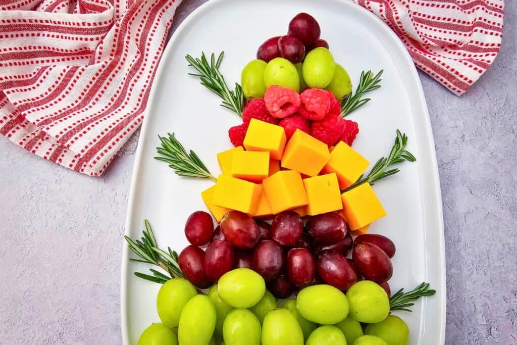 A fruit Christmas tree arranged on a white oval platter with layers of green and red grapes, fresh raspberries, cheddar cheese cubes, and rosemary sprigs, framed by a red and white striped holiday towel.