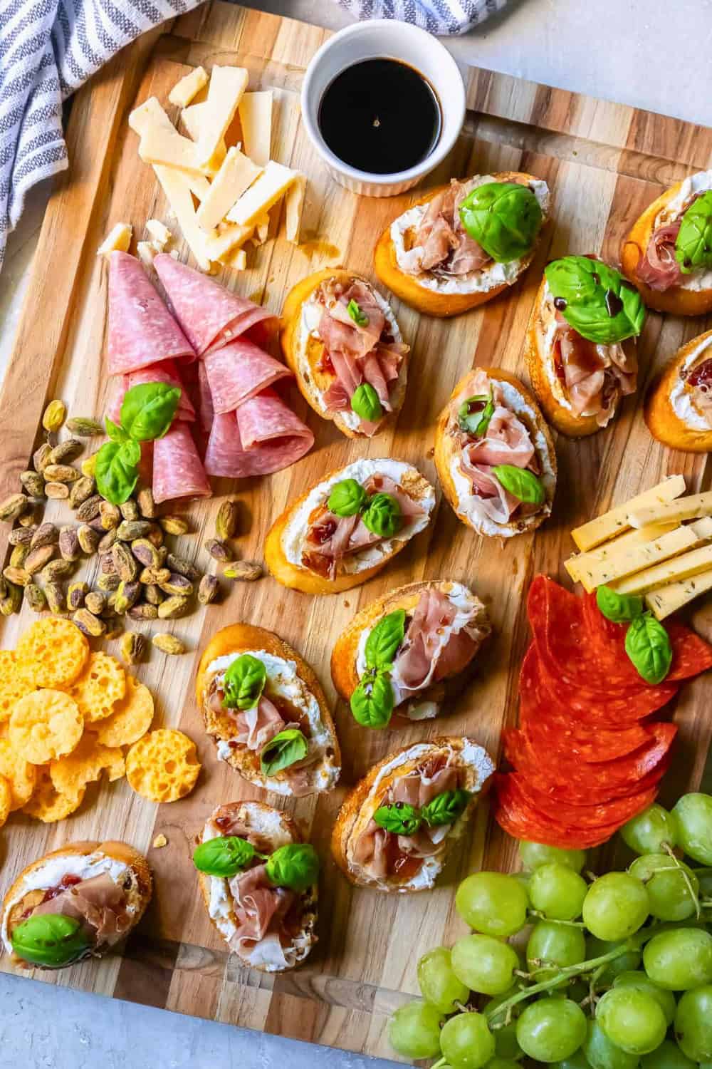 A wooden appetizer board with goat cheese, fig and prosciutto crostini topped with fresh basil and balsamic glaze, surrounded by salami, pepperoni, cheeses, pistachios, crackers and green grapes for a festive Christmas party potluck appetizer.