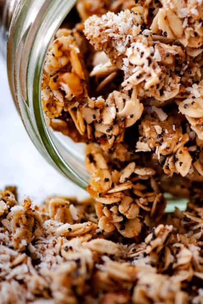 Close up of lemon poppy seed granola spilling from a glass jar, showing toasted oats, coconut flakes, chopped nuts, and tiny black poppy seeds throughout.