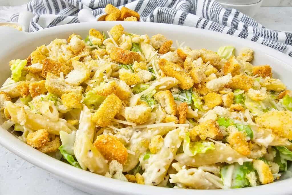 Creamy Caesar pasta salad in a white serving dish topped with crunchy croutons, shaved Parmesan, and chopped romaine lettuce, styled as a rich and creamy pasta salad ready to serve.