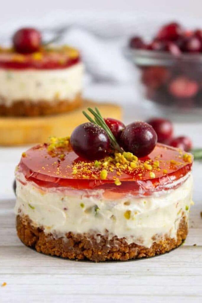 Mini pistachio cranberry cheesecake with a graham cracker crust, creamy filling speckled with pistachio pieces, and a glossy cranberry jelly topping. Garnished with sugared cranberries, crushed pistachios, and a sprig of rosemary for a festive holiday look.