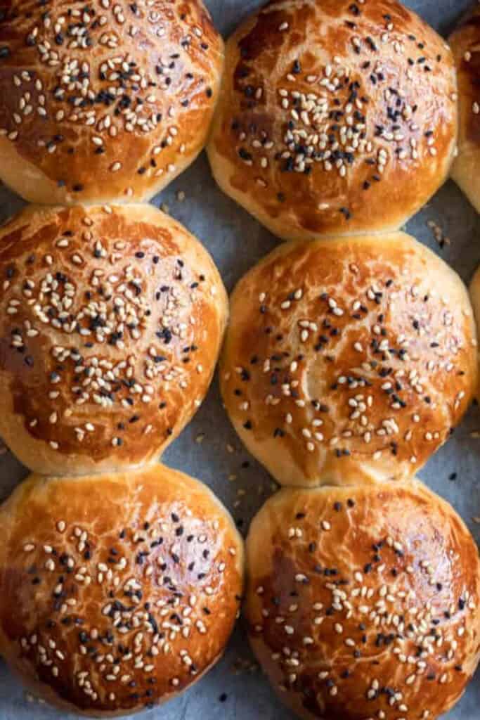 reshly baked Turkish cheese buns arranged on a baking tray with glossy golden brown tops sprinkled with white and black sesame seeds. The soft homemade rolls are shown close up, making them an appealing road trip snack for kids and teens.