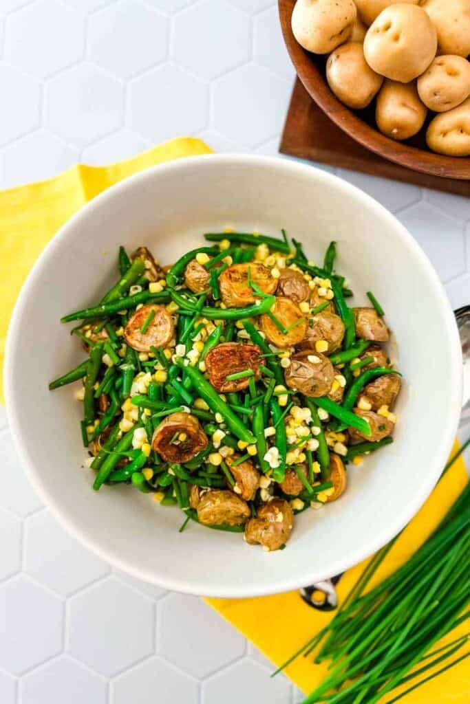 A fresh potato and green bean salad served in a white bowl, featuring roasted baby potatoes, crisp green beans, and sweet corn kernels, garnished with chopped chives. The dish sits on a bright yellow napkin, with a wooden bowl of raw baby potatoes nearby for a rustic touch.