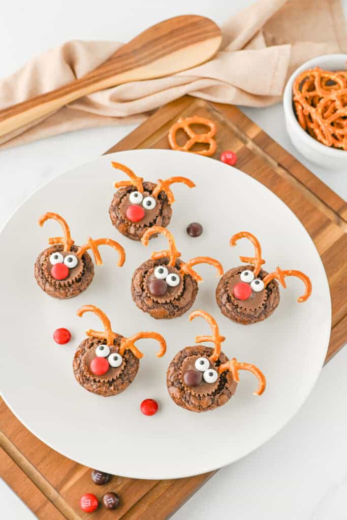 A white plate filled with reindeer brownie bites decorated with chocolate peanut butter cups, candy eyes, red and brown candy noses, and broken pretzels shaped like antlers.
