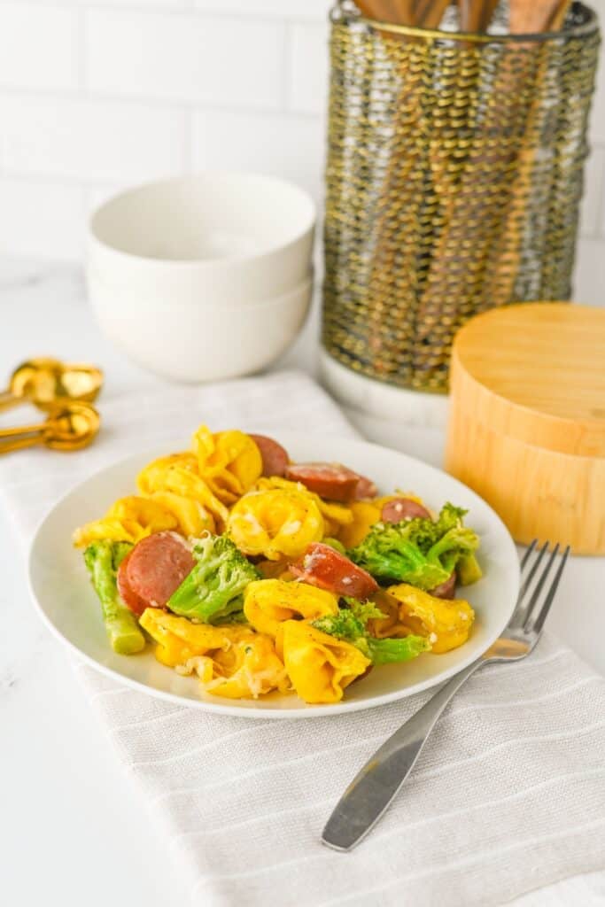 Plate of sausage, tortellini, and broccoli resting on a striped kitchen towel, with a cozy kitchen setup in the background. This sausage veggies sheet pan meal is vibrant and cheesy, combining smoked sausage, tender pasta, and roasted vegetables for a quick and satisfying dinner.