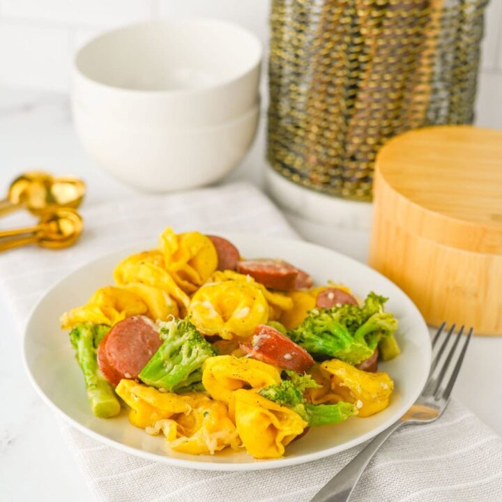 Plate of sausage, tortellini, and broccoli resting on a striped kitchen towel, with a cozy kitchen setup in the background. This sausage veggies sheet pan meal is vibrant and cheesy, combining smoked sausage, tender pasta, and roasted vegetables for a quick and satisfying dinner.