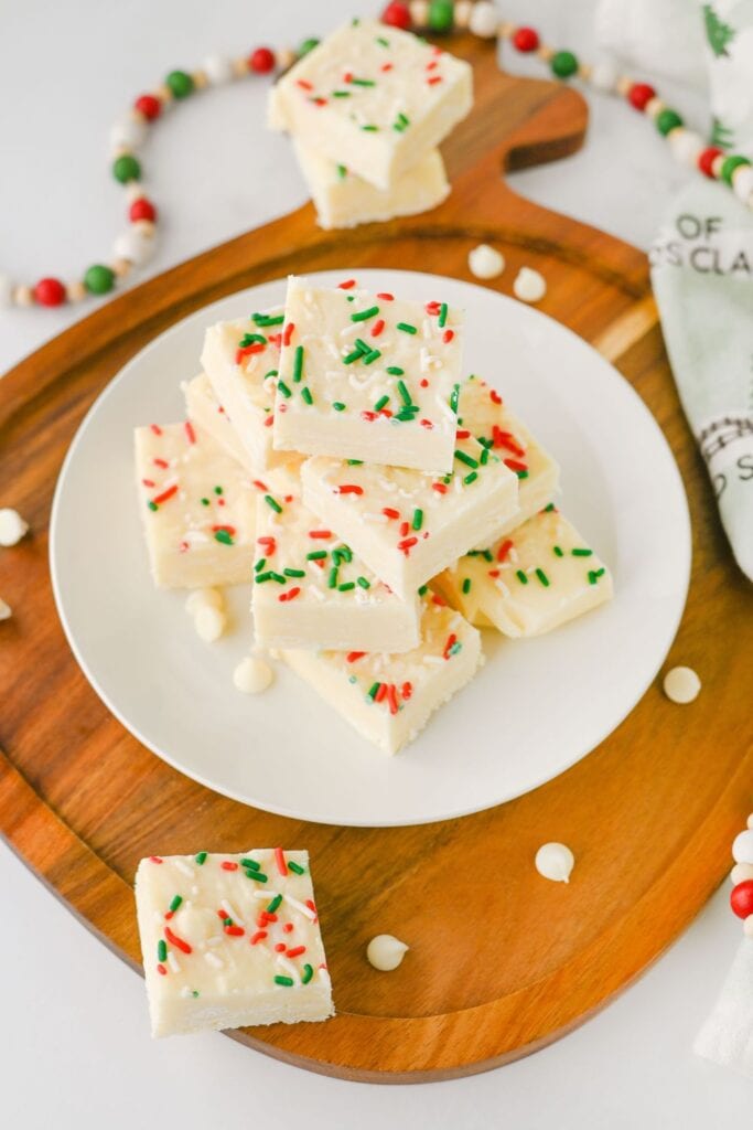Stack of white chocolate fudge squares on a white plate, topped with festive red, green, and white sprinkles. The easy shortcut Christmas candy treat is displayed on a wooden board with scattered white chocolate chips and holiday decorations.