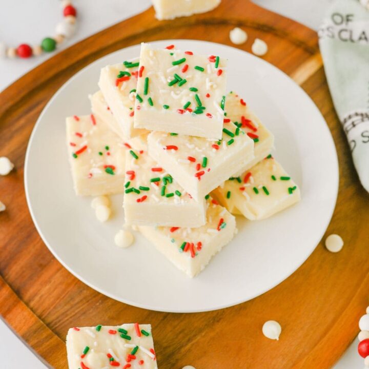 Stack of white chocolate fudge squares on a white plate, topped with festive red, green, and white sprinkles. The easy shortcut Christmas candy treat is displayed on a wooden board with scattered white chocolate chips and holiday decorations.